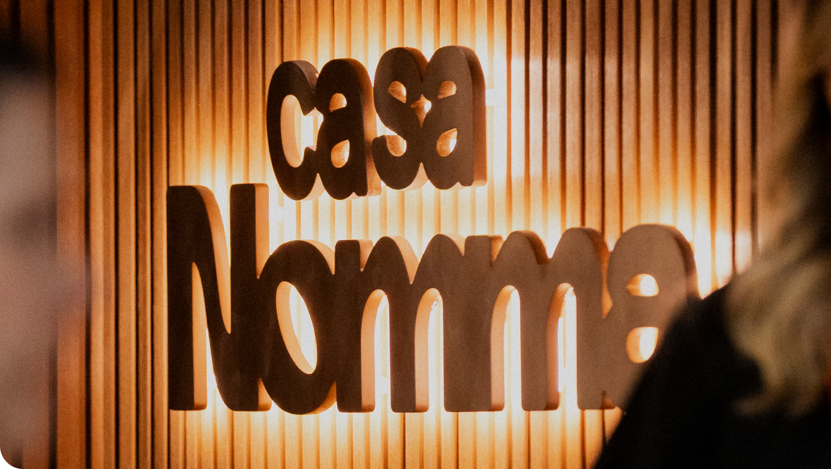 Letreiramento da marca Casa Nomma em relevo sobre painel ripado de madeira iluminado.