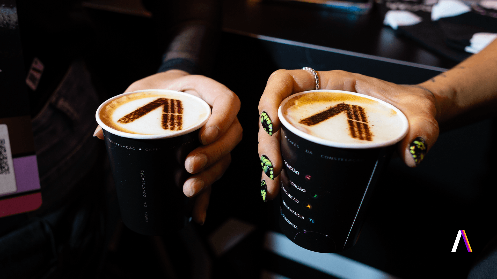 Café da Constelação: duas mãos seguram copos com o logo da Layer Up desenhado na espuma, com os copos estampados com as palavras conexão, exploração, projeção, ressonância e conquista